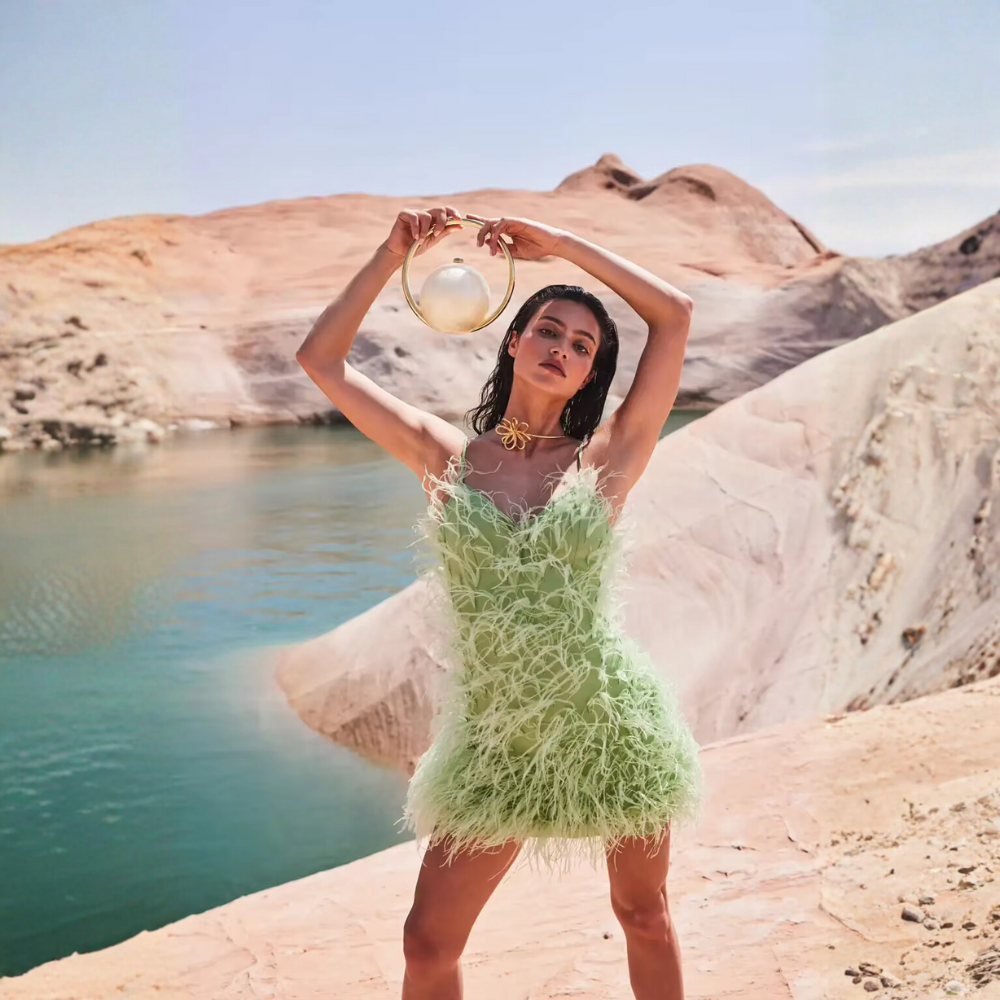 “Woman in a light green dress holding a marble acrylic clutch near a blue lake and sandy cliffs.”